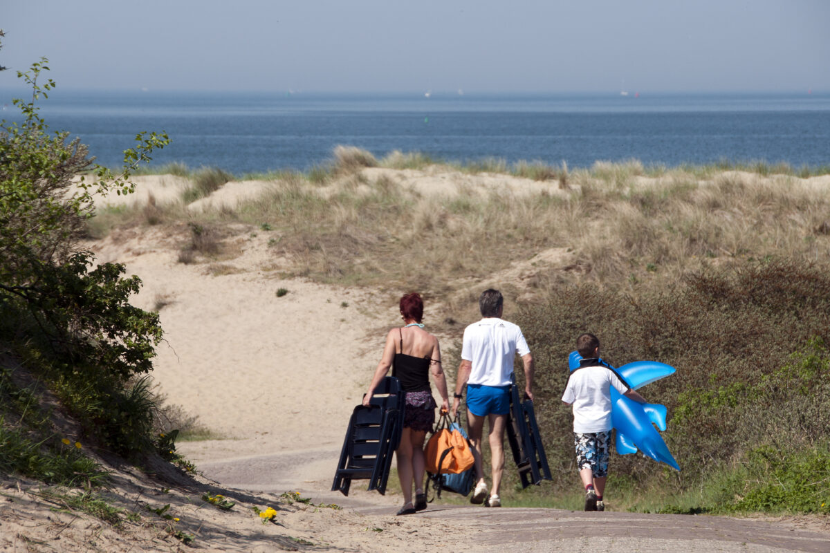 Gezin loopt via een duinpad richting het strand, met strandstoelen en een opblaasdier in de hand.