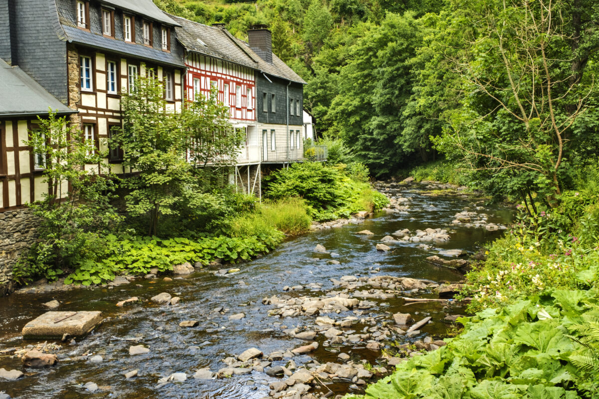 Het vakwerkstadje Monschau in de Eifel, Duitsland, met gekleurde huizen langs de rivier de Rur en groene heuvels op de achtergrond.