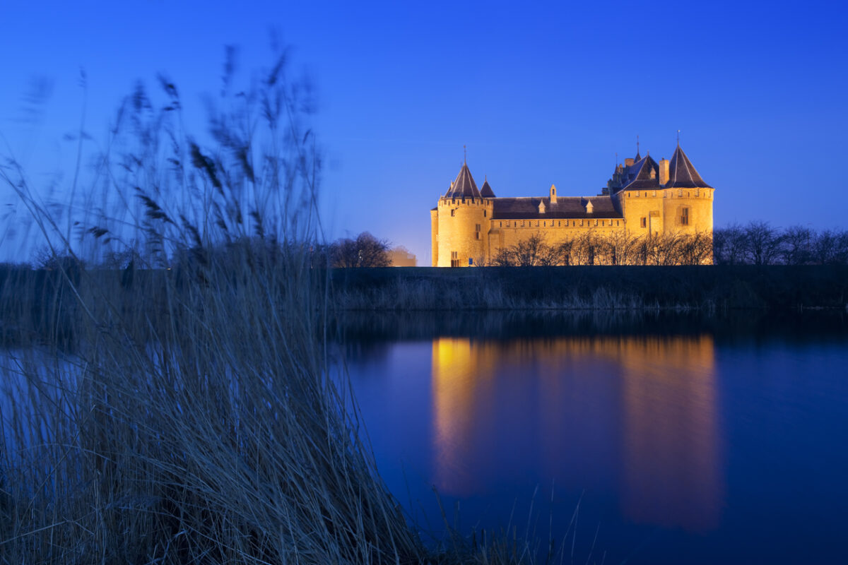 Muiderslot bij avondlicht, weerspiegeld in het water onder een diepblauwe hemel.